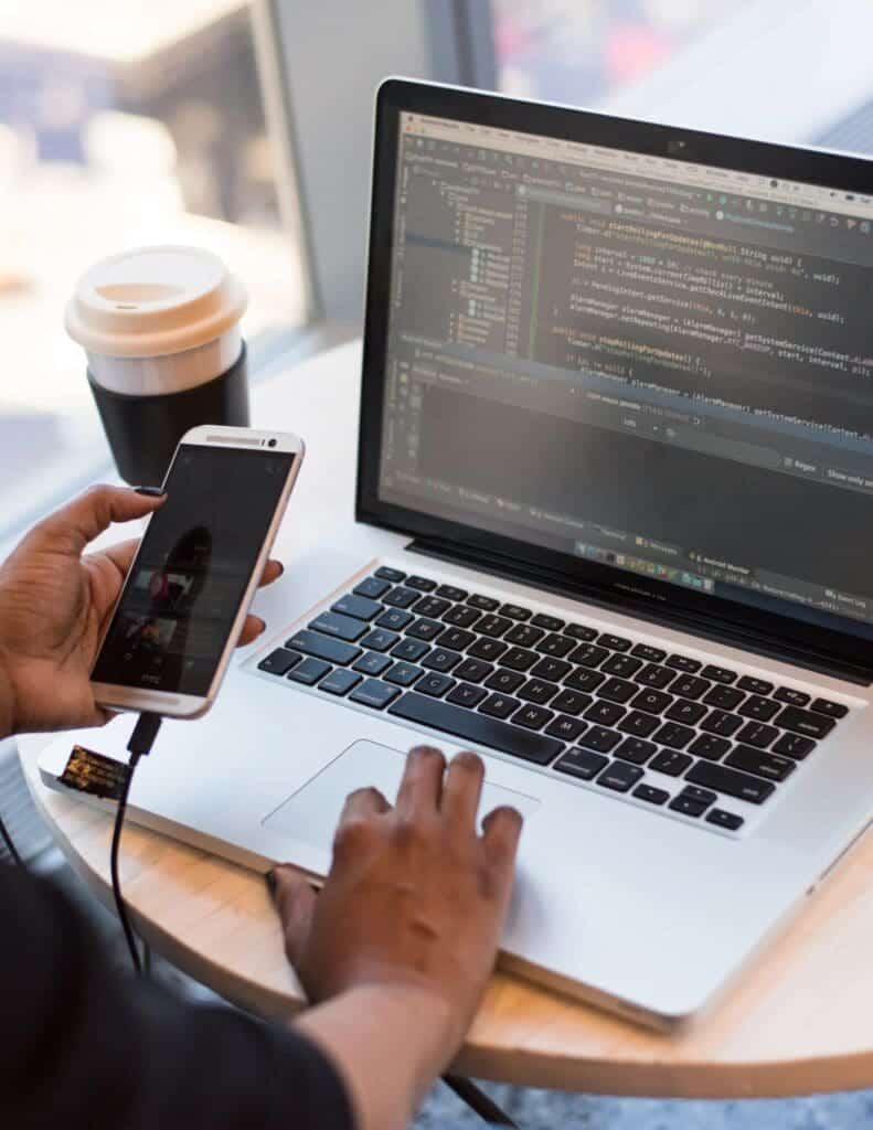 A pair of hands. One hand holds a smartphone while the other sits on the trackpad of a laptop showing code.