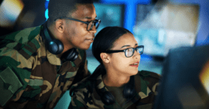 A man and a woman in military uniforms and glasses smile as they look at a computer screen.