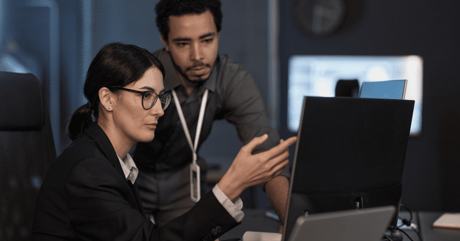 A woman in a suit and glasses gestures to a computer monitor while a man in a gray button-down looks over her shoulder.