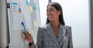 A woman in a gray plaid blazer uses a dry erase marker to gesture to a chart pinned to a whiteboard.