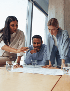 Three young professionals wearing business casual attire lean over a table looking at a large sheet of paper.