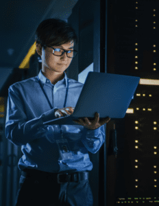 A man stands in a dark server room and looks at the laptop he is holding.