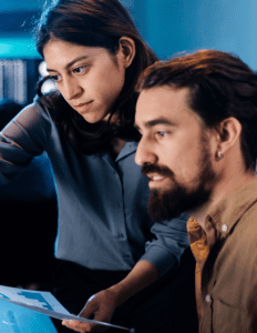 A man and woman look at a computer off camera while the woman holds a piece of paper with a graph on it.