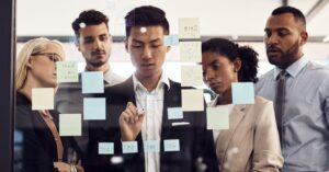 Five people wearing business attire look at post-it notes while one writes using a dry erase marker.