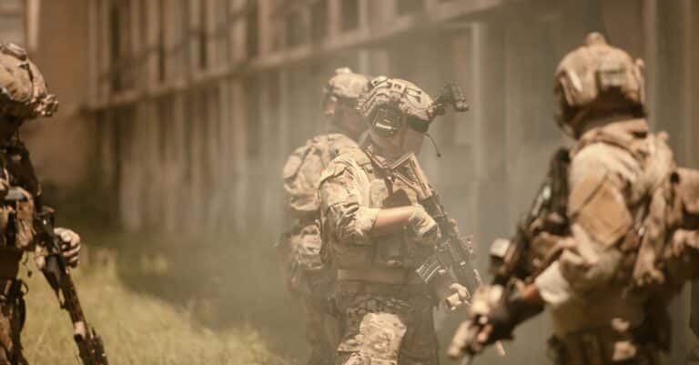 Soldiers in tan camouflage uniforms hold guns next to a fence.