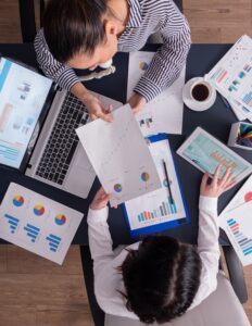 Two people sit at a table with coffee, a laptop, and papers showing graphs on it.