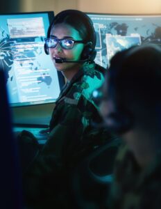 A woman in a military uniform and headset turns around in her seat in front of a computer monitor.