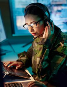 A woman in a military uniform and glasses holds a headset to her ear and looks at a computer monitor.