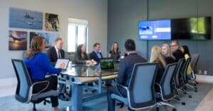 People in business attire talk while sitting around a glass table in desk chairs.