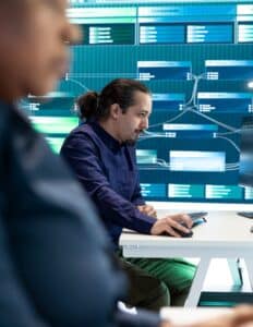 A man in a blue button down uses a mouse while looking at a desktop monitor. A large screen is behind him.