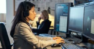 A woman wearing a tan blazer and headphones types on a computer keyboard.