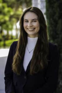 Professional headshot of Sydney Wright, a smiling woman with long brown hair, wearing a white blouse and dark blazer, standing outdoors with a blurred green background.