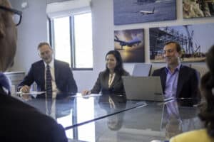 A professional business meeting with three individuals seated at a glass conference table, engaged in discussion. The background features aviation and construction-themed artwork.