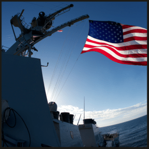 An American flag waving on a U.S. Navy ship with a clear blue sky and ocean in the background.