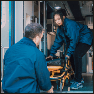 Two emergency medical responders in navy blue uniforms loading a stretcher into an ambulance.