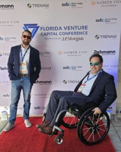 Two men pose on a red carpet at the Florida Venture Capital Conference. The man on the left, wearing a blazer, jeans, and sunglasses, stands with a relaxed posture. The man on the right, wearing a suit and sunglasses, sits in a wheelchair while popping a wheelie. The event backdrop behind them displays sponsor logos, including J.P. Morgan, Silicon Valley Bank, and Trenam Law.