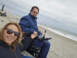 A smiling woman wearing sunglasses takes a selfie while holding hands with a man in a power wheelchair on a sandy beach. The man, dressed in a blue jacket, holds a cup and looks at the camera with ocean waves rolling in behind him under an overcast sky.