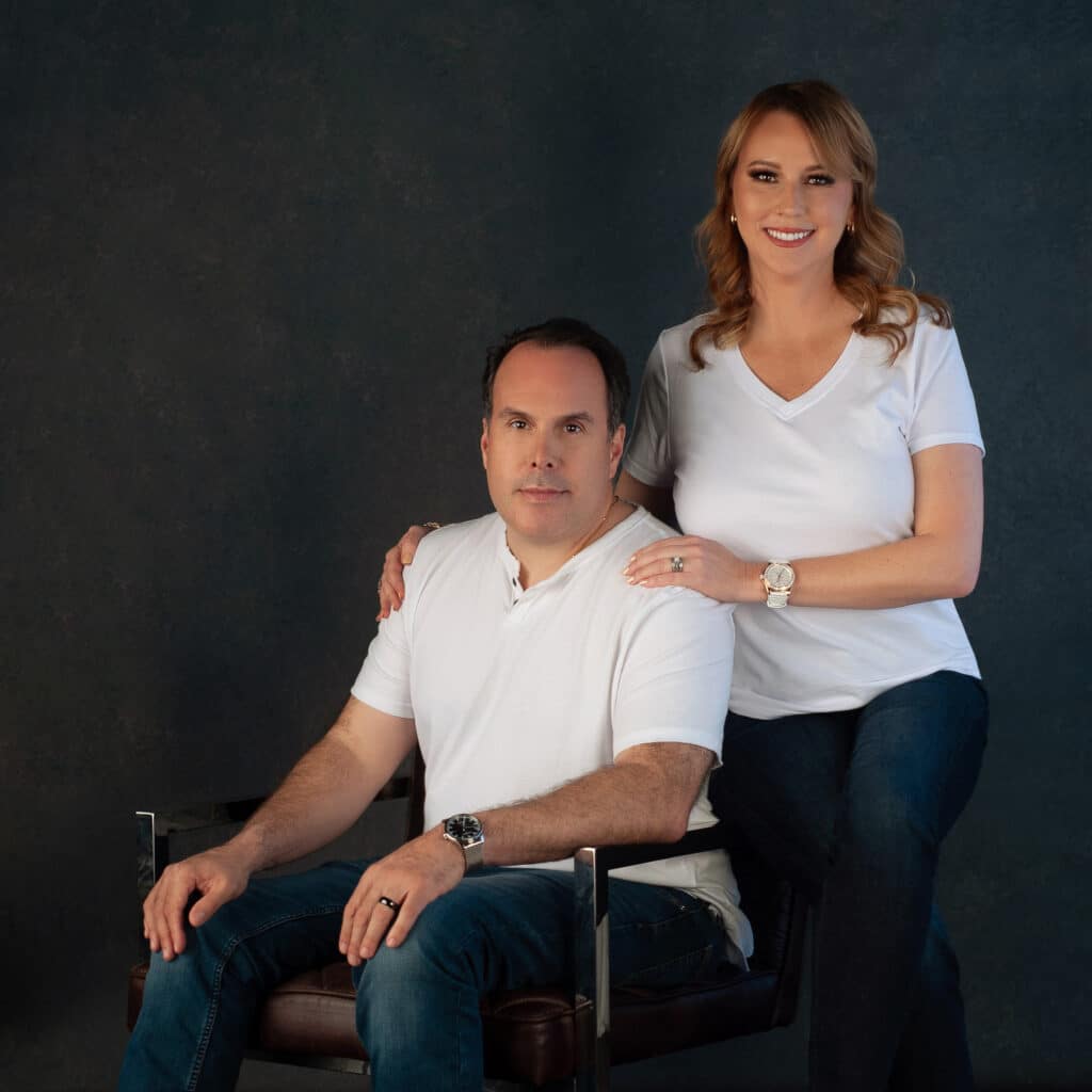 A man and a woman in matching white t-shirts and jeans pose for a professional portrait. The man sits in a leather chair, while the woman stands beside him with her hand on his shoulder, smiling. The background is dark and textured.