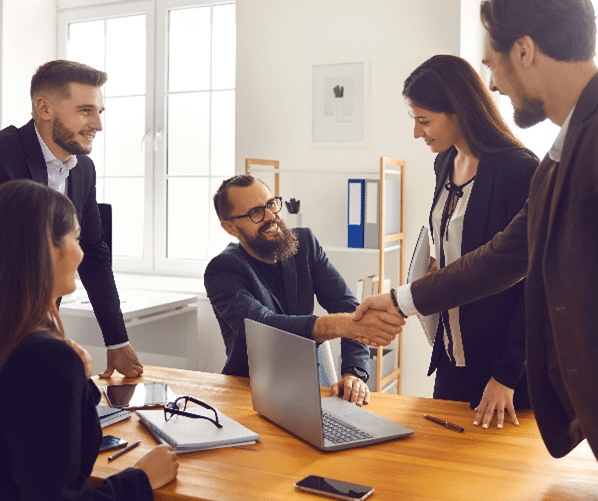 A group of business men and women shaking hands over a wooden table and laptop.