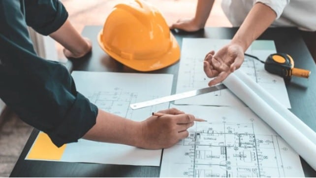 Two people collaborate on architectural blueprints at a table, surrounded by a yellow hard hat and measuring tools, conveying teamwork and planning.