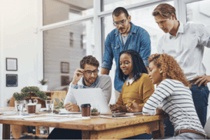 A diverse group of five colleagues collaborates around a laptop in a bright office. Their focused expressions convey teamwork and engagement.