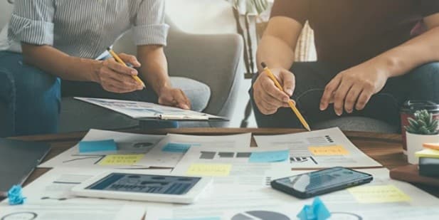 Two people sit at a table with scattered documents, smartphones, and sticky notes. They hold pencils, seemingly engaged in a collaborative discussion.