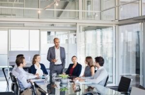 A diverse group of six professionals sit around a glass table in a modern conference room. One person stands, speaking confidently. The group listens attentively.