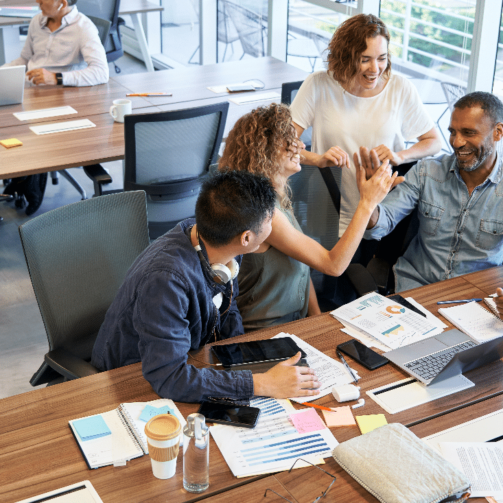 A group of professional colleagues smiling and congratulating each other at a wooden table scattered with work documents.