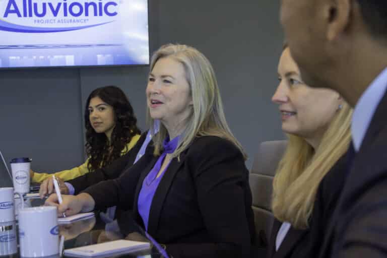 A group of professionals sitting at a conference table during a meeting at Alluvionic, with a company logo visible on the screen in the background.