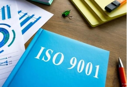 A blue book on a desk with papers. The book has white lettering that says ISO 9001.