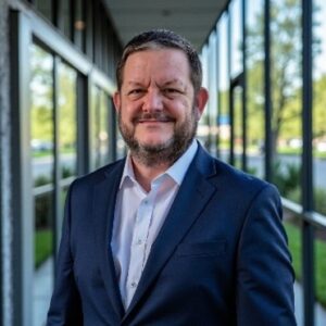 Portrait of Mike Crandall in a navy suit, standing outdoors in front of a glass building corridor.