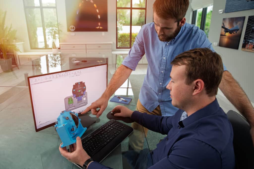 Two engineers review a 3D CAD model on a computer while one holds a blue 3D-printed mechanical component and the other points to the screen.