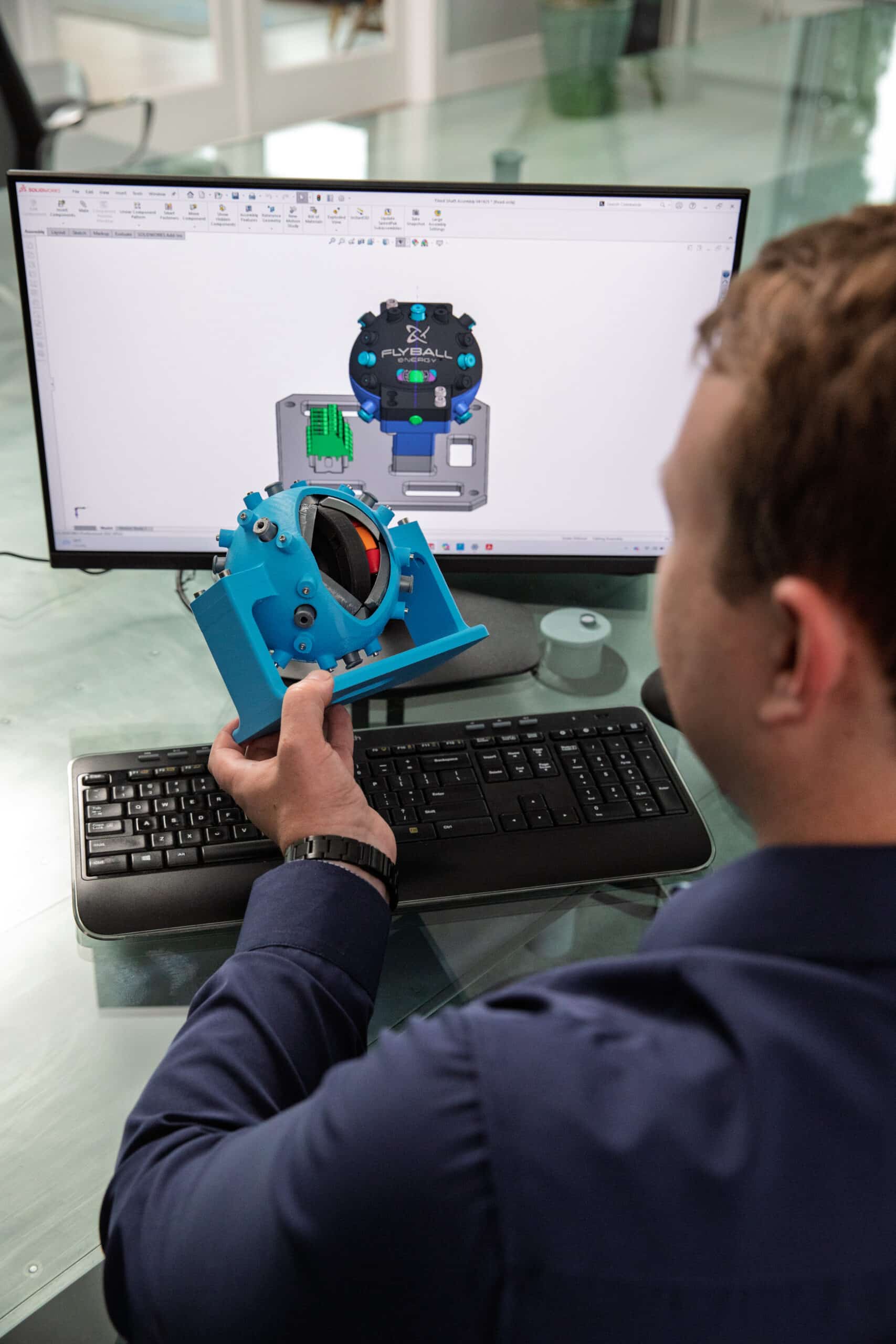 A professional holding a blue mechanical component while comparing it to a 3D design displayed on a computer monitor in a modern office.