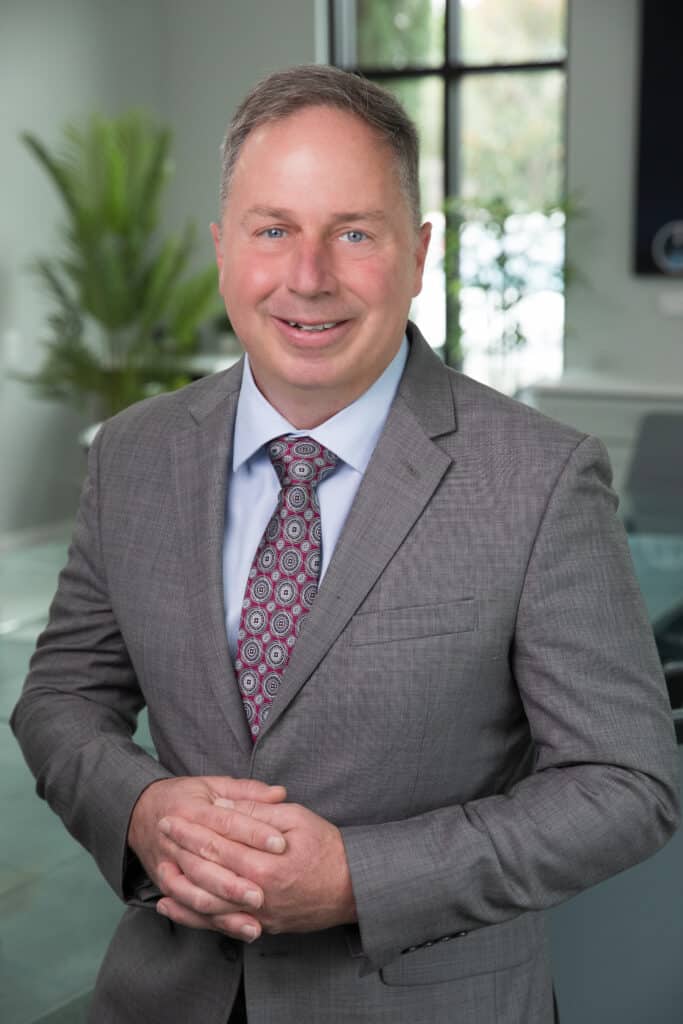 Corporate portrait of a man in a gray suit and patterned tie, standing in a modern office.