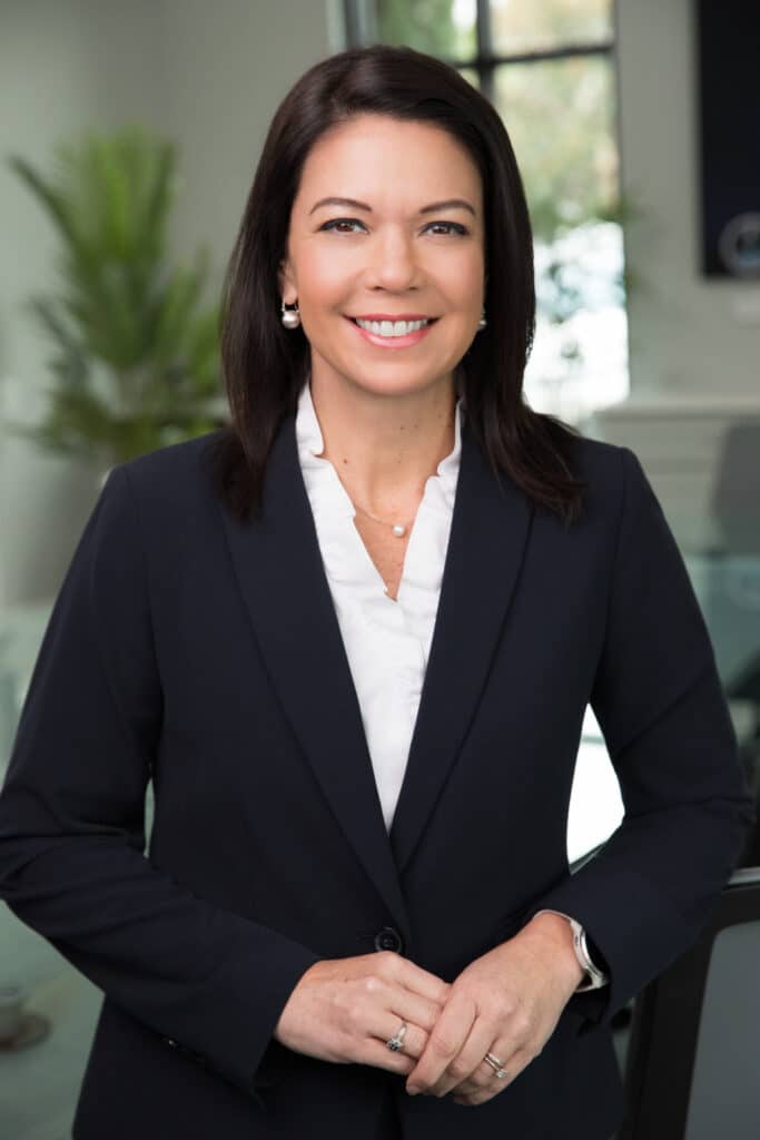 Corporate headshot of a woman in a dark blazer and white blouse, smiling in an office environment.