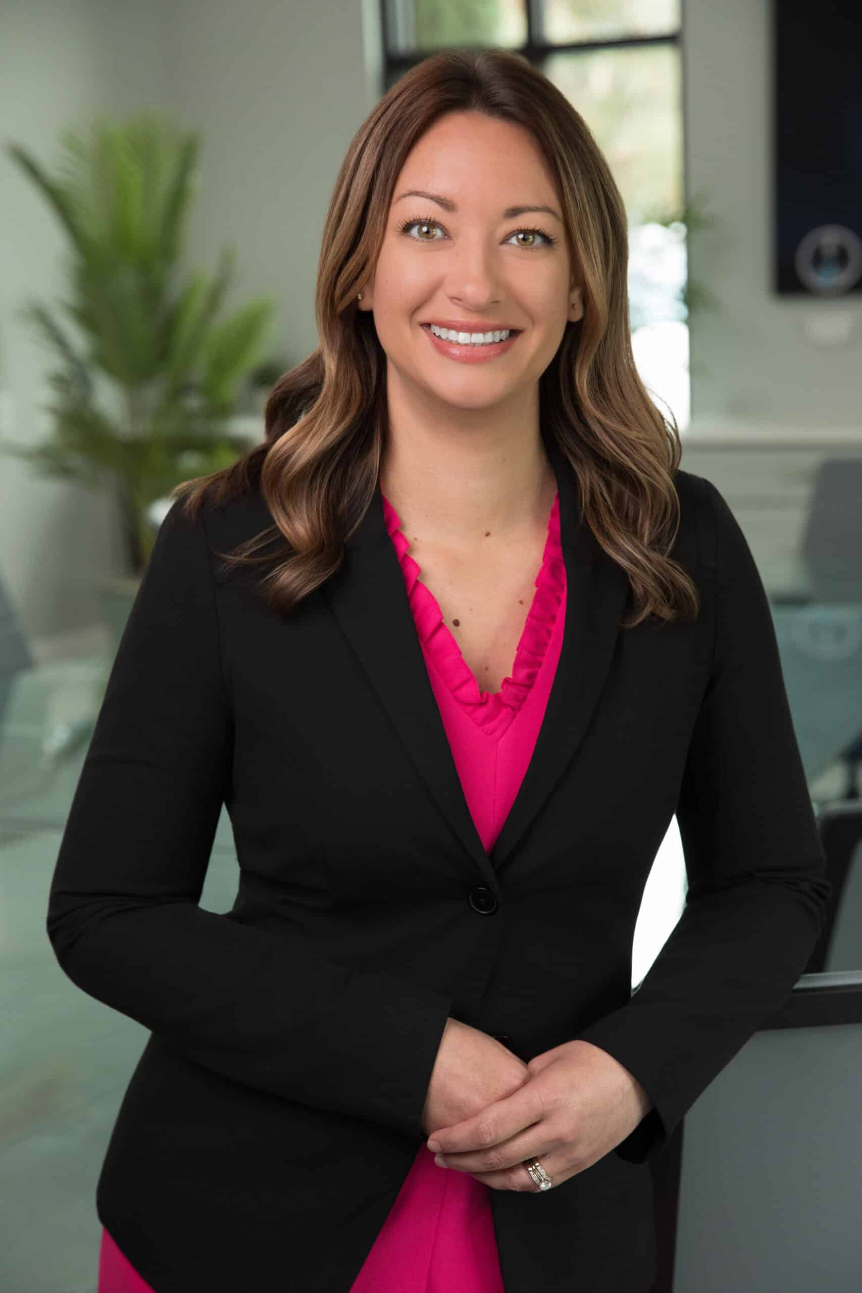 Professional portrait of a woman in a black blazer and bright pink blouse, standing in an office.