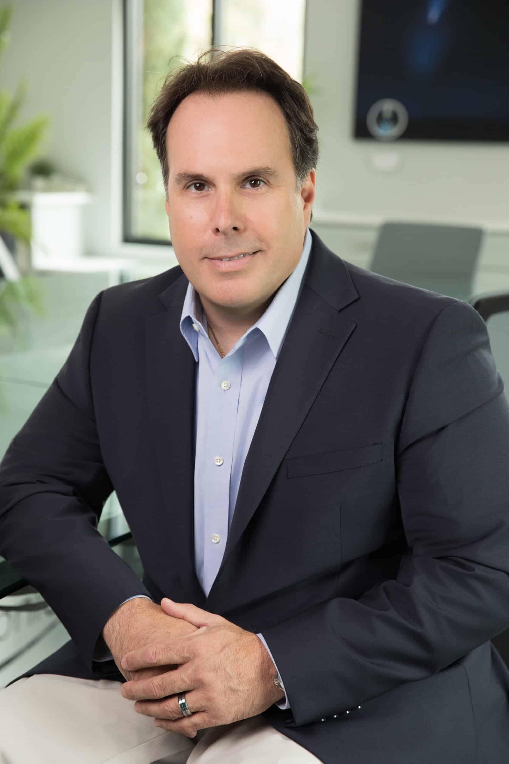 Professional headshot of a man in a dark blazer and gray shirt, leaning forward in an office.