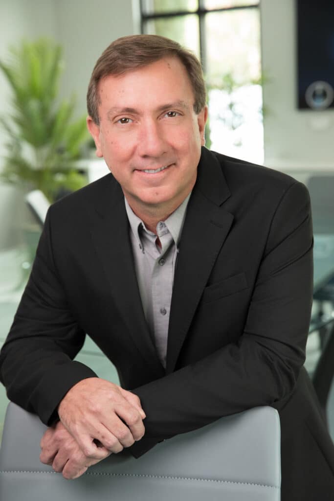 Professional headshot of a man in a navy blazer and light blue shirt, seated in an office.