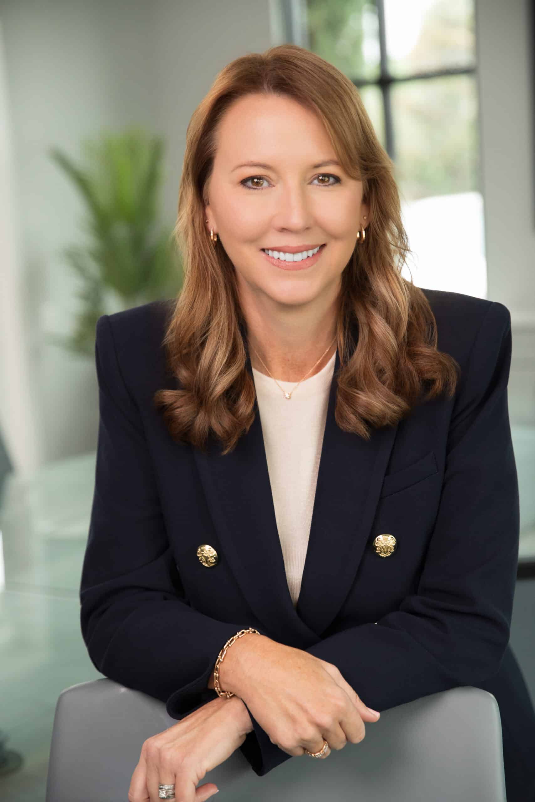 Professional headshot of a woman in a navy blazer, smiling while seated in a modern office.