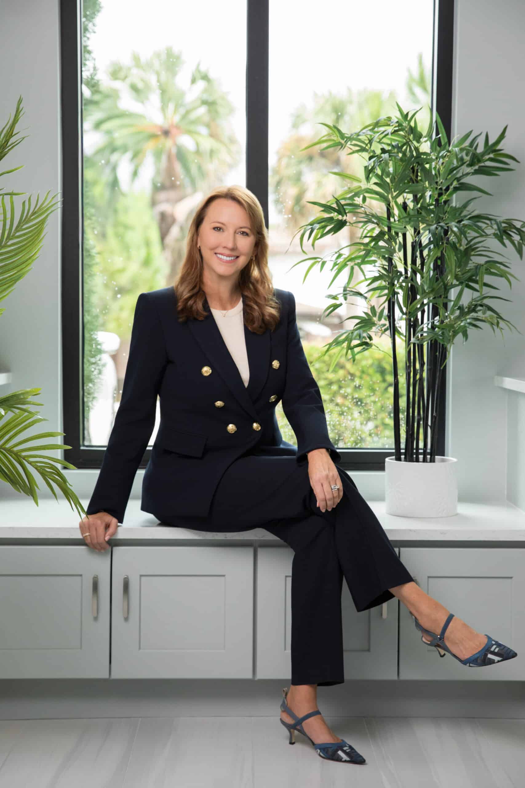 Professional portrait of a woman in a navy suit seated on a built-in bench by a large window, smiling in a bright, modern office with plants.